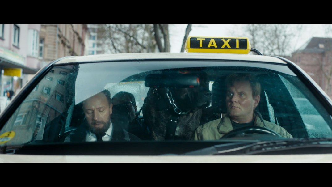 Ein Taxi mit zwei Männern und einem großen Hund auf der Rückbank steht auf einer Stadtstraße. Der Fahrer schaut nachdenklich, während der Beifahrer schläft., © Barnsteiner-Film/Across Nations Ein Taxi mit zwei Männern und einem großen Hund auf der Rückbank steht auf einer Stadtstraße. Der Fahrer schaut nachdenklich, während der Beifahrer schläft., © Barnsteiner-Film/Across Nations