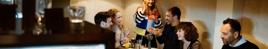 A group of people are sitting around a table in a café and laughing. A woman stands and talks to the others. The atmosphere is cozy., © Stuttgart Marketing, Alwin Maigler