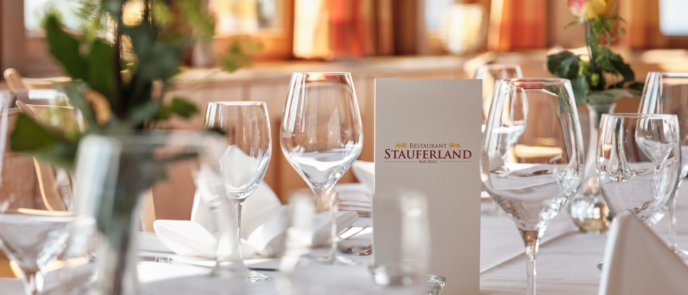 Eleganter Tisch im Restaurant Stauferland, gedeckt mit Weingläsern, Besteck und Blumen. Im Hintergrund sind große Fenster zu sehen., © Badhotel Restaurant Stauferland