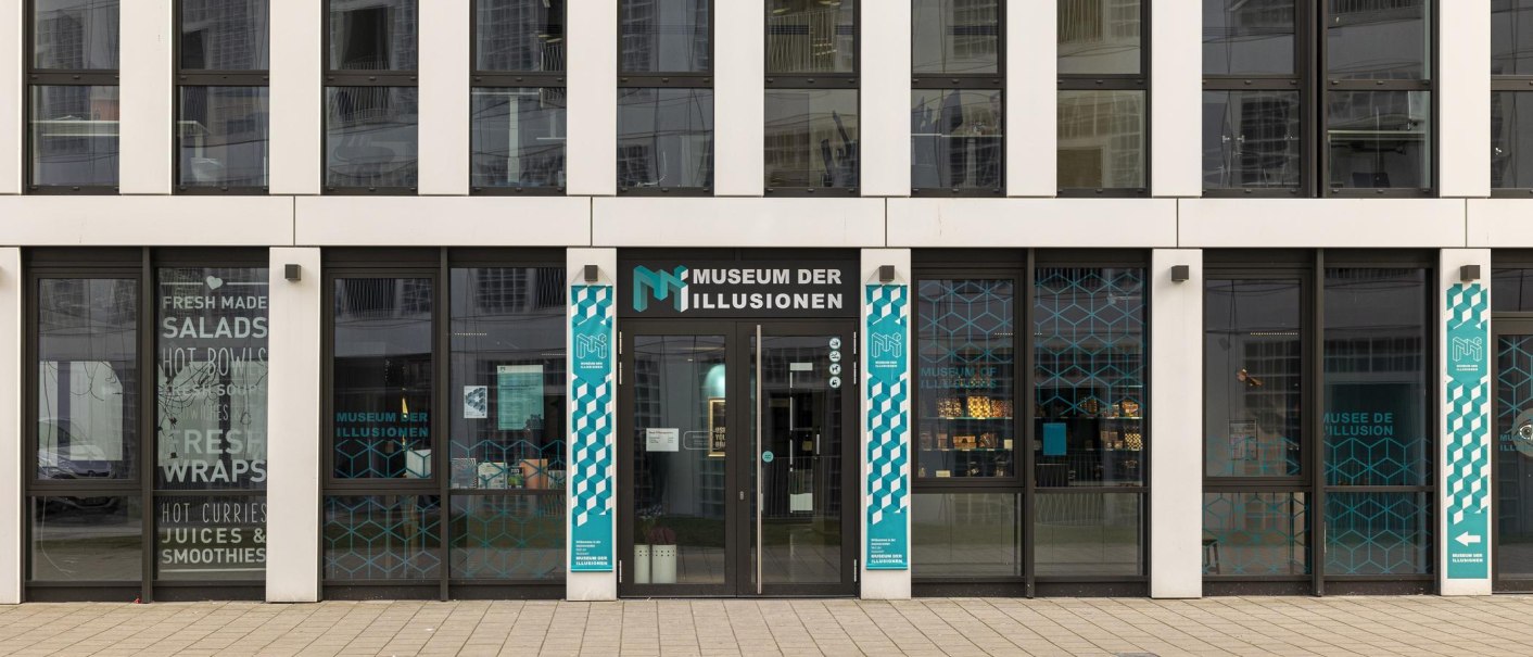 Eingang des Museums der Illusionen in Stuttgart. Moderne Glasfassade mit türkisfarbenen Grafiken und Schriftzug. Schilder weisen auf das Museum hin., © Stuttgart-Marketing GmbH, Sarah Schmid