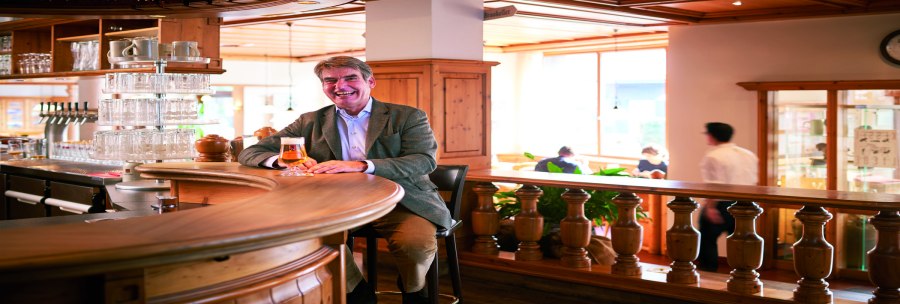 A man sits smiling at a wooden bar in a brewery pub, holding a glass of beer. Guests and a waiter can be seen in the background., © © Stuttgart-Marketing GmbH