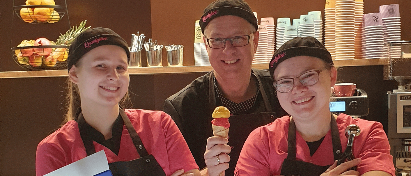 Three people in an ice cream parlor smile at the camera. One person is holding an ice cream, with colorful cups and fruit in the background., &copy; kesselglueck.de