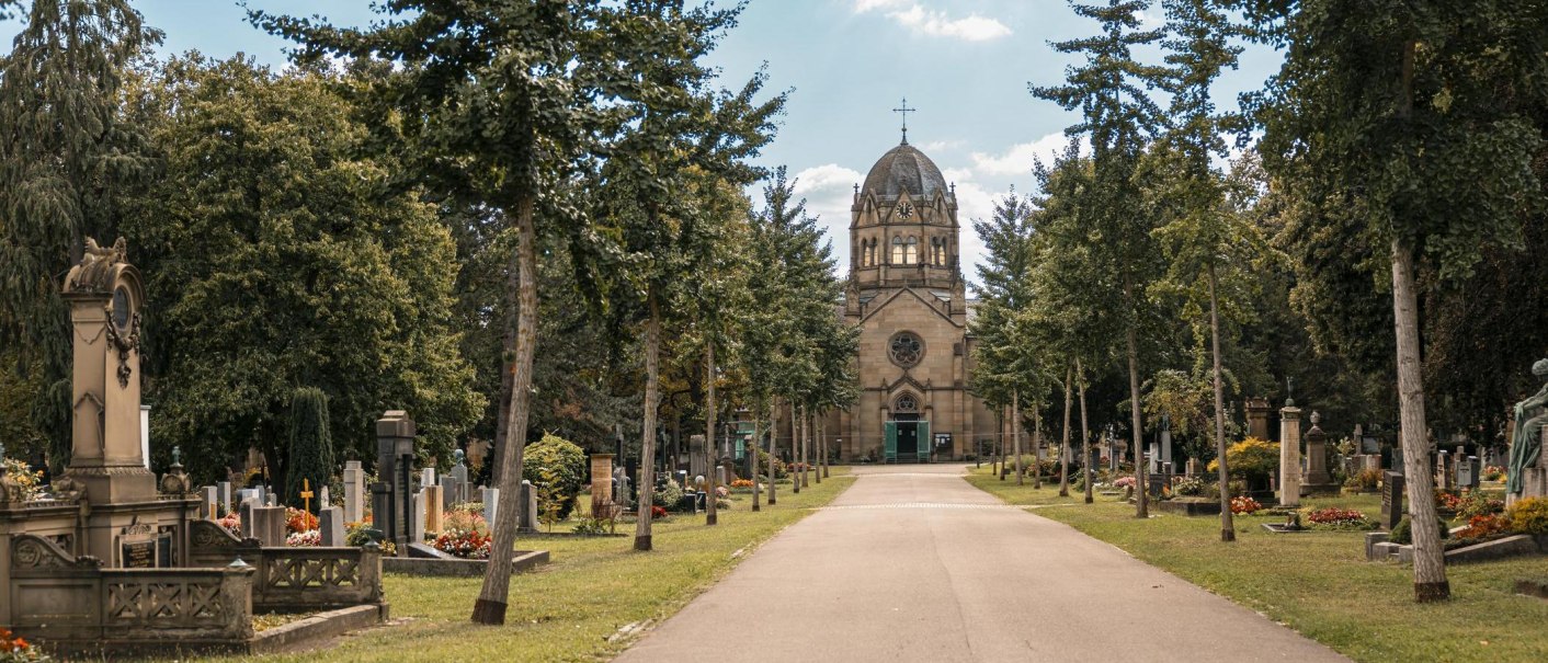 Ein Weg führt durch einen Friedhof mit Bäumen und Grabsteinen, im Hintergrund eine Kirche mit Kuppel und Kreuz., © Stuttgart Marketing GmbH, Sarah Schmid