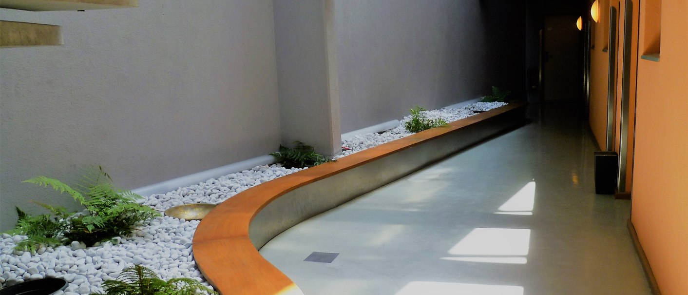 Moderner Flur mit geschwungenem Pflanzbeet, weißen Kieselsteinen und Farnen. Orange Wand mit runden Lampen, Wegweiser zur Rezeption sichtbar., © Abalon Hotel Ideal Moderner Flur mit geschwungenem Pflanzbeet, weißen Kieselsteinen und Farnen. Orange Wand mit runden Lampen, Wegweiser zur Rezeption sichtbar., © Abalon Hotel Ideal