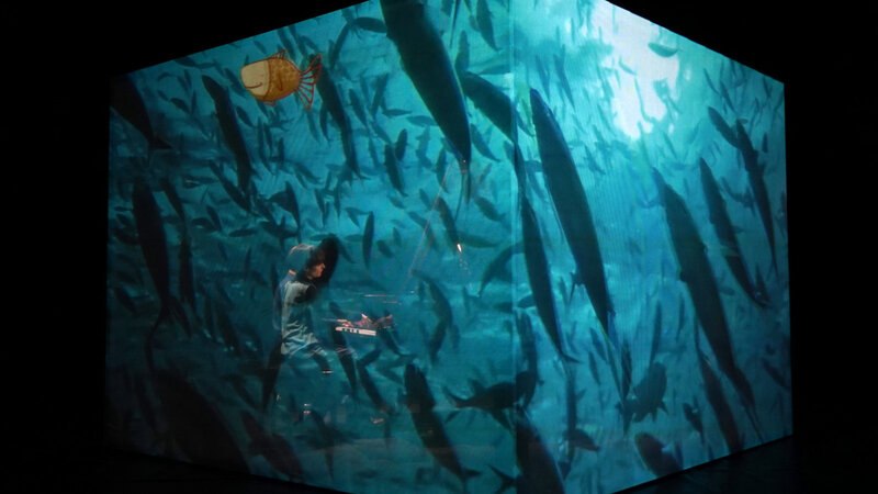 Ein Raum mit Projektionen von Fischen, in dem ein Musiker Klavier spielt. Die Atmosphäre ist aquatisch und künstlerisch., © FITZ Das Theater animierter Formen Ein Raum mit Projektionen von Fischen, in dem ein Musiker Klavier spielt. Die Atmosphäre ist aquatisch und künstlerisch., © FITZ Das Theater animierter Formen