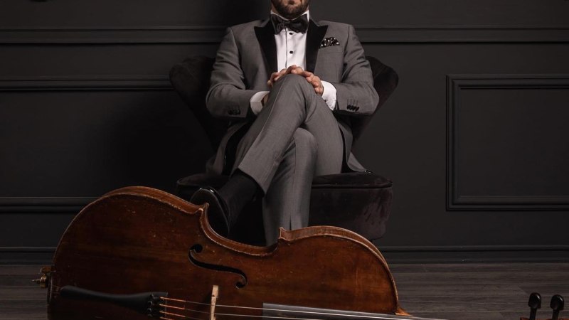 Ein elegant gekleideter Mann im grauen Anzug sitzt vor einer dunklen Wand. Ein Cello liegt vor ihm auf dem Boden., &copy; Elem&eacute;r Duka