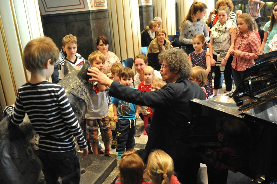 Ein Mann interagiert mit einer Gruppe von Kindern in einem Raum mit einem Klavier. Die Kinder sind aufmerksam und neugierig., © STUTTGARTER SALONIKER Ein Mann interagiert mit einer Gruppe von Kindern in einem Raum mit einem Klavier. Die Kinder sind aufmerksam und neugierig., © STUTTGARTER SALONIKER