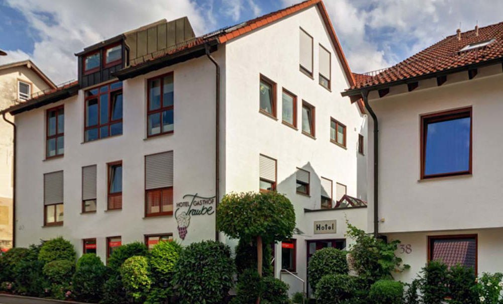 Weißes Hotelgebäude mit roten Fenstern, umgeben von grünen Büschen. Der Himmel ist bewölkt., © TOMAS Weißes Hotelgebäude mit roten Fenstern, umgeben von grünen Büschen. Der Himmel ist bewölkt., © TOMAS
