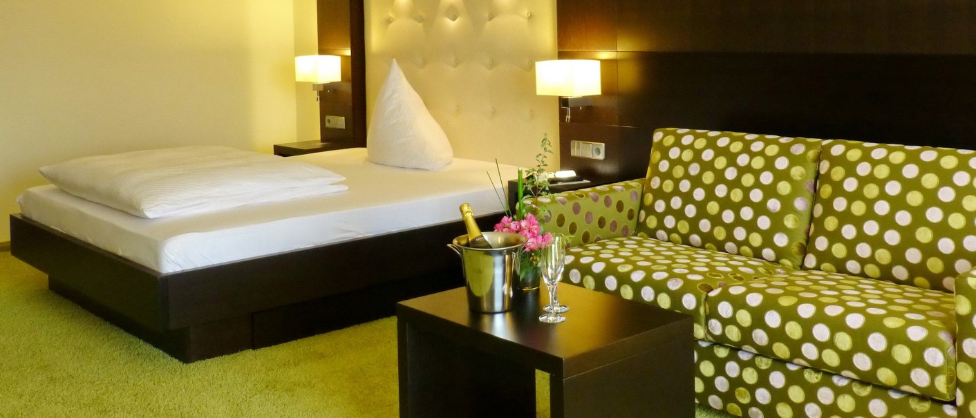 Modern hotel room with double bed, green dotted sofa, table with champagne cooler and glasses, on a green carpeted floor., © Hotel & Restaurant Sonne