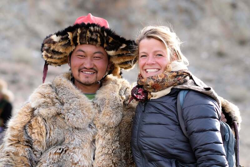A man in traditional fur clothing and a woman in modern winter clothing smile at the camera. They are standing in front of a blurred background outdoors., &copy; Theaterhaus Stuttgart e.V.
