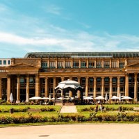 The Königsbau in Stuttgart with its classicist façade and glass roof, surrounded by green spaces and walkers in sunny weather., © Stuttgart-Marketing GmbH, Sarah Schmid
