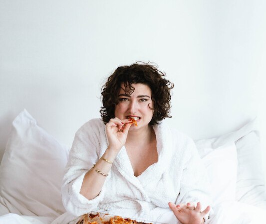 Eine Person im weißen Bademantel sitzt im Bett und isst Lasagne aus einer Auflaufform., © Rosenau Kultur e.V. Eine Person im weißen Bademantel sitzt im Bett und isst Lasagne aus einer Auflaufform., © Rosenau Kultur e.V.