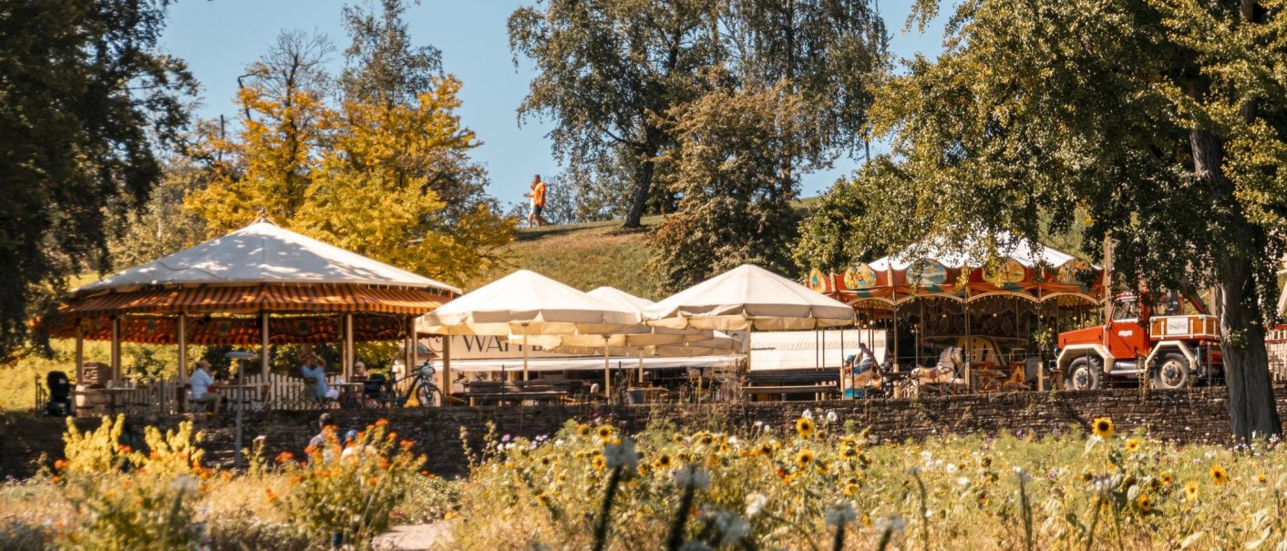 Ein Jahrmarkt im Killesbergpark mit Karussell, Sonnenschirmen und blühender Wiese im Vordergrund. Menschen genießen das sonnige Wetter., © Stuttgart-Marketing GmbH, Sarah Schmid