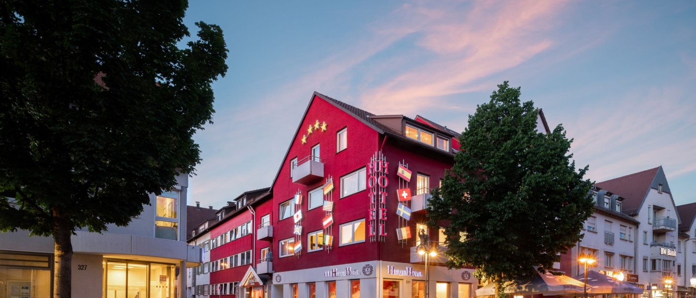 Rotes Hotelgebäude mit Flaggen und beleuchteten Schildern bei Dämmerung, umgeben von Bäumen und Straßenlaternen., © Hetzel Hotel Stuttgart Rotes Hotelgebäude mit Flaggen und beleuchteten Schildern bei Dämmerung, umgeben von Bäumen und Straßenlaternen., © Hetzel Hotel Stuttgart