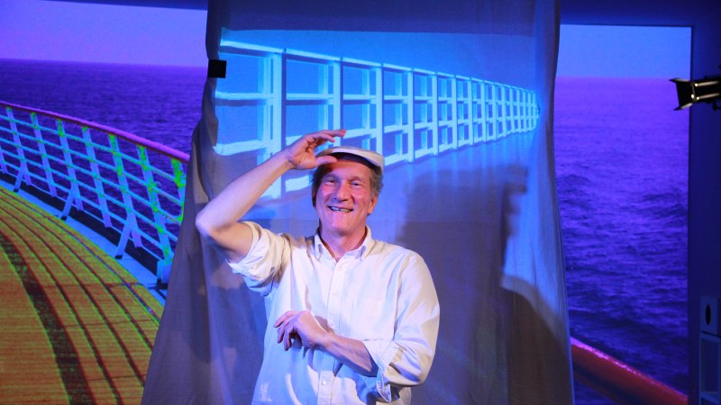 Ein Mann in wei&szlig;em Hemd und Hut sitzt l&auml;chelnd vor einer Leinwand, die ein Schiffsdeck zeigt. Die Szene erinnert an eine Theaterauff&uuml;hrung., &copy; Katja Ritter