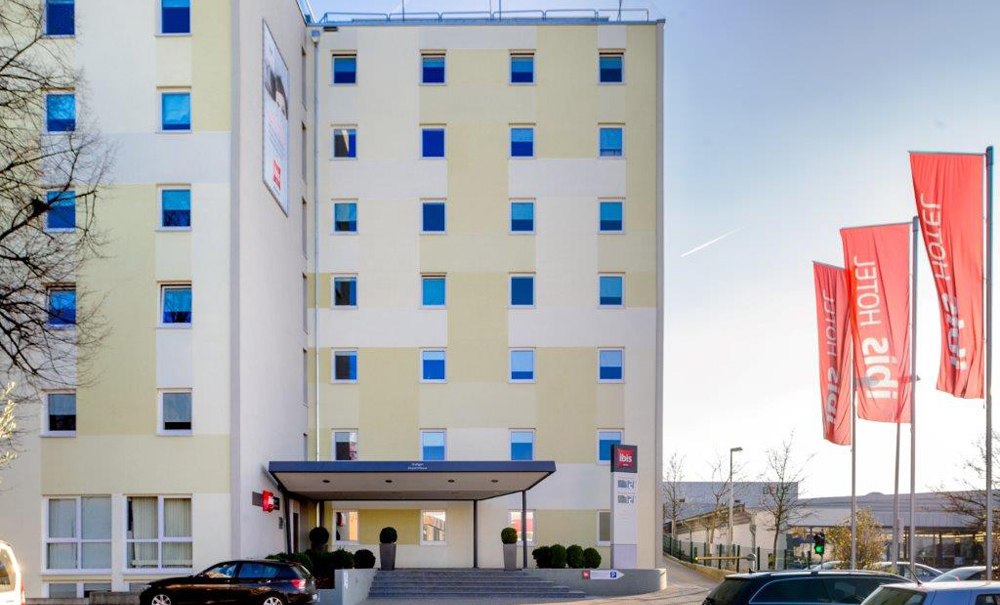 Das Ibis Hotel Stuttgart Airport Messe mit roten Fahnen und geparkten Autos davor. Das Gebäude hat mehrere Stockwerke und eine helle Fassade., © Ibis Stuttgart Airport Messe