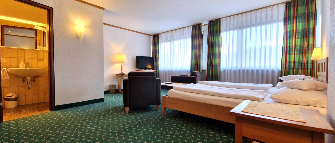 Spacious hotel room with a double bed, an armchair, a TV, and an adjoining bathroom. Large windows with checkered curtains let in plenty of light., © TOMAS Spacious hotel room with a double bed, an armchair, a TV, and an adjoining bathroom. Large windows with checkered curtains let in plenty of light., © TOMAS