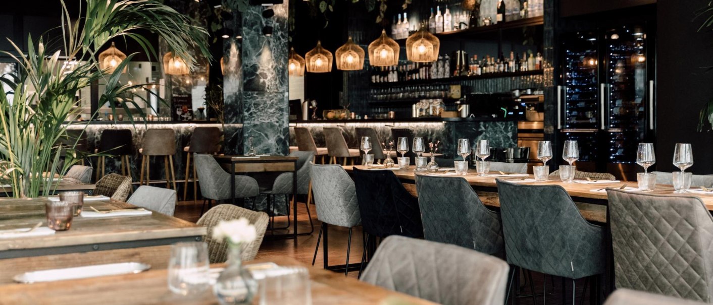 Elegant restaurant with modern furniture, laid tables and a stylish bar in the background. Subtle lighting and plants create a cozy atmosphere., © Malo Stuttgart - Lea Prestle