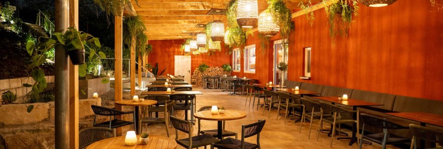 Cozy outdoor area with wooden tables, chairs and hanging plants. Warm lighting and red wall create an inviting atmosphere., &copy; SatzDrei