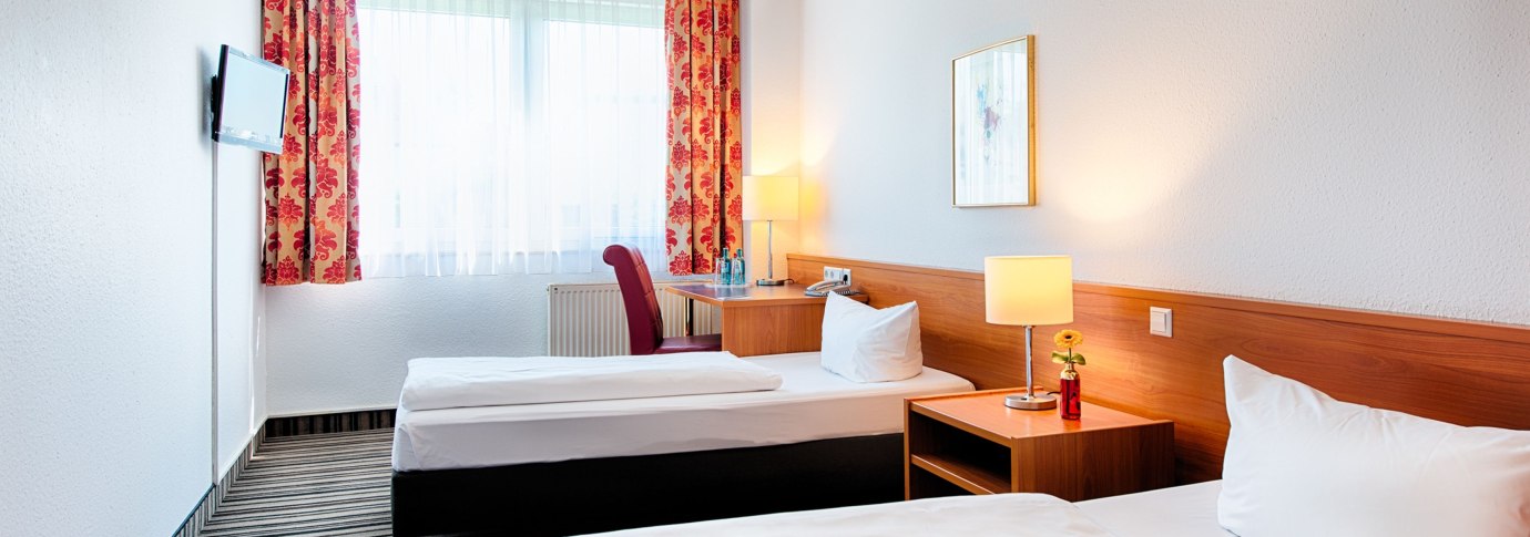 Helles Zweibettzimmer mit zwei Einzelbetten, Nachttisch mit Lampe, Schreibtisch, Fernseher und roten Vorh&auml;ngen., &copy; ACHAT Hotels Deutschland