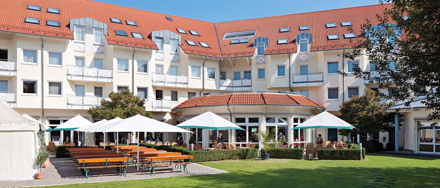 Biergarten mit Holzbänken und weißen Sonnenschirmen vor einem großen Gebäude mit roten Dächern und vielen Fenstern., © Seminaris Hotel Bad Boll