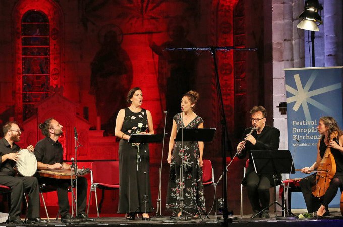 Musiker und Sänger auf einer Bühne, beleuchtet in Rot, bei einem Kirchenmusikfestival. Ein Banner mit der Aufschrift 'Festival Europäischer Kirchenmusik' ist sichtbar., © Landesmuseum Württemberg