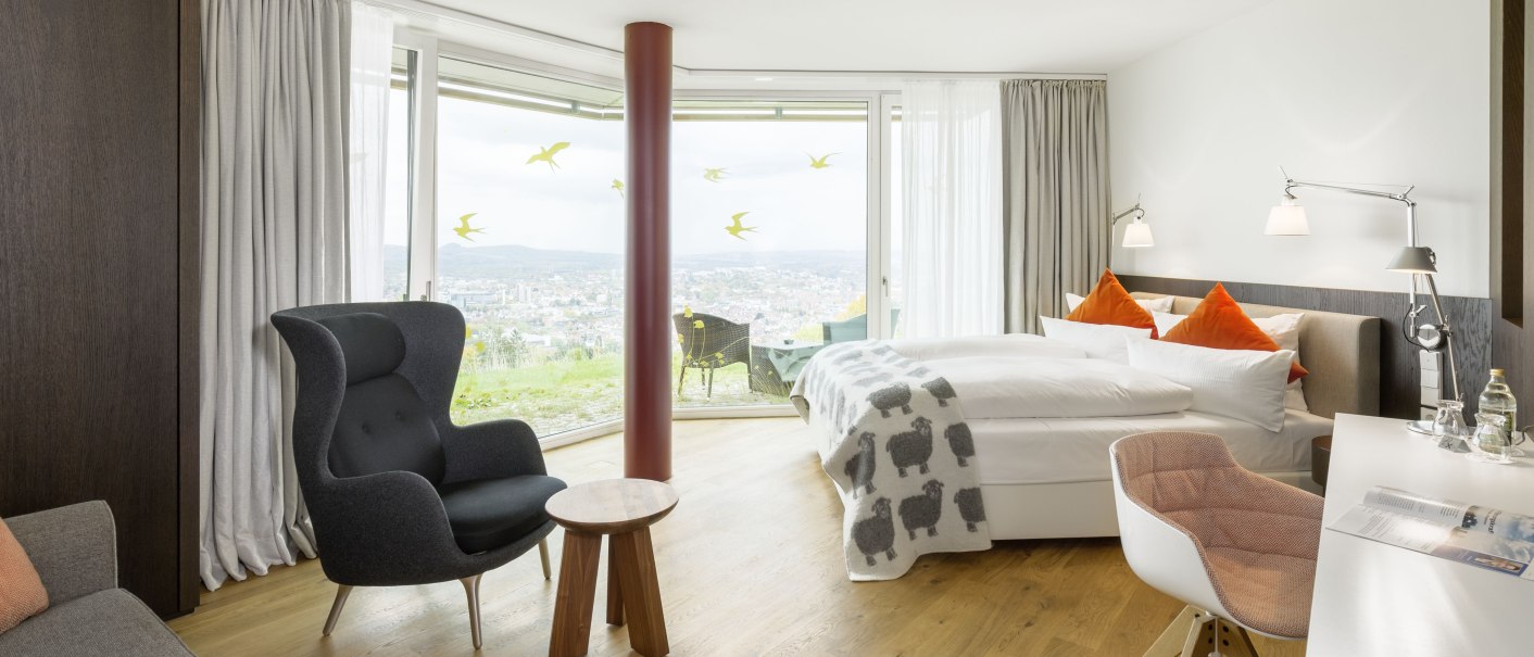 Moderne Hotelsuite mit großem Fenster, gemütlichem Sessel, Schreibtisch und Doppelbett. Blick auf die Stadtlandschaft durch das Fenster., © Achalm. Hotel GmbH & Co. KG Moderne Hotelsuite mit großem Fenster, gemütlichem Sessel, Schreibtisch und Doppelbett. Blick auf die Stadtlandschaft durch das Fenster., © Achalm. Hotel GmbH & Co. KG