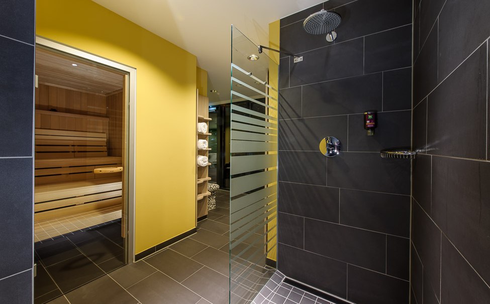 Moderne Dusche mit dunklen Fliesen, Glaswand und Regendusche. Im Hintergrund ist eine Sauna mit Holzbänken und gelber Wand zu sehen., © TOMAS Moderne Dusche mit dunklen Fliesen, Glaswand und Regendusche. Im Hintergrund ist eine Sauna mit Holzbänken und gelber Wand zu sehen., © TOMAS