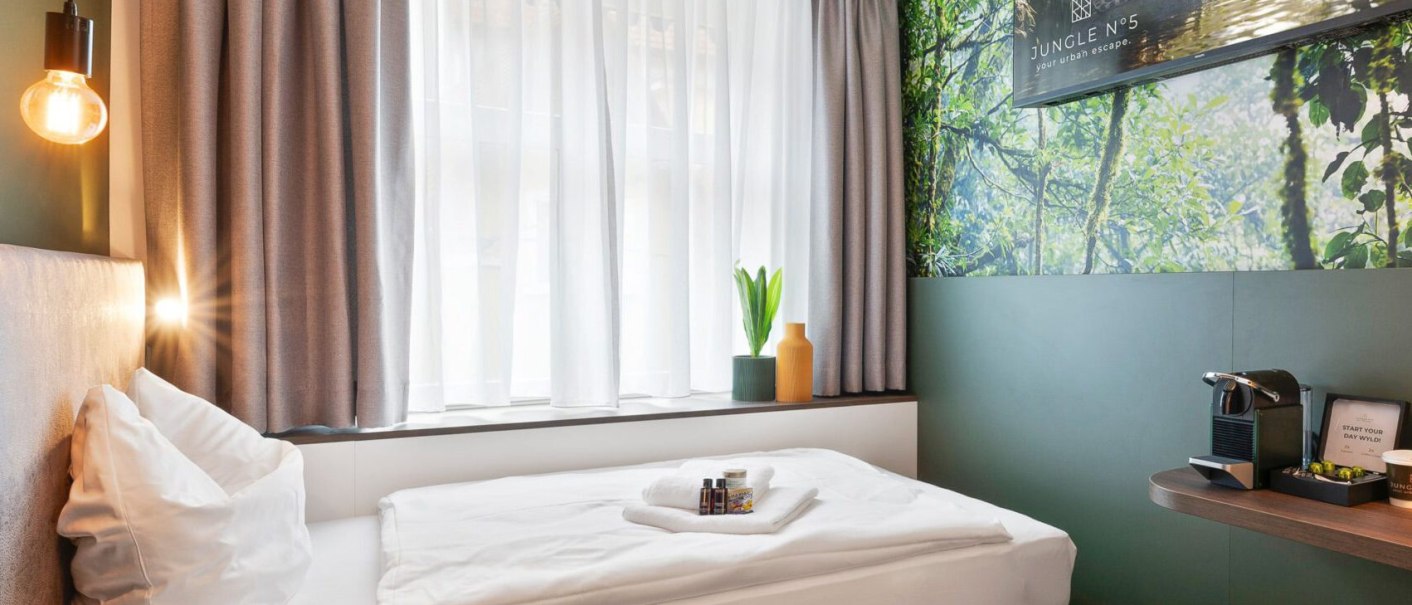 Modernes Einzelzimmer mit Bett, gro&szlig;em Fenster, Dschungel-Wandbild und Kaffeemaschine. Dezente Beleuchtung und stilvolle Dekoration., &copy; JUNGLE N&deg;5 GmbH