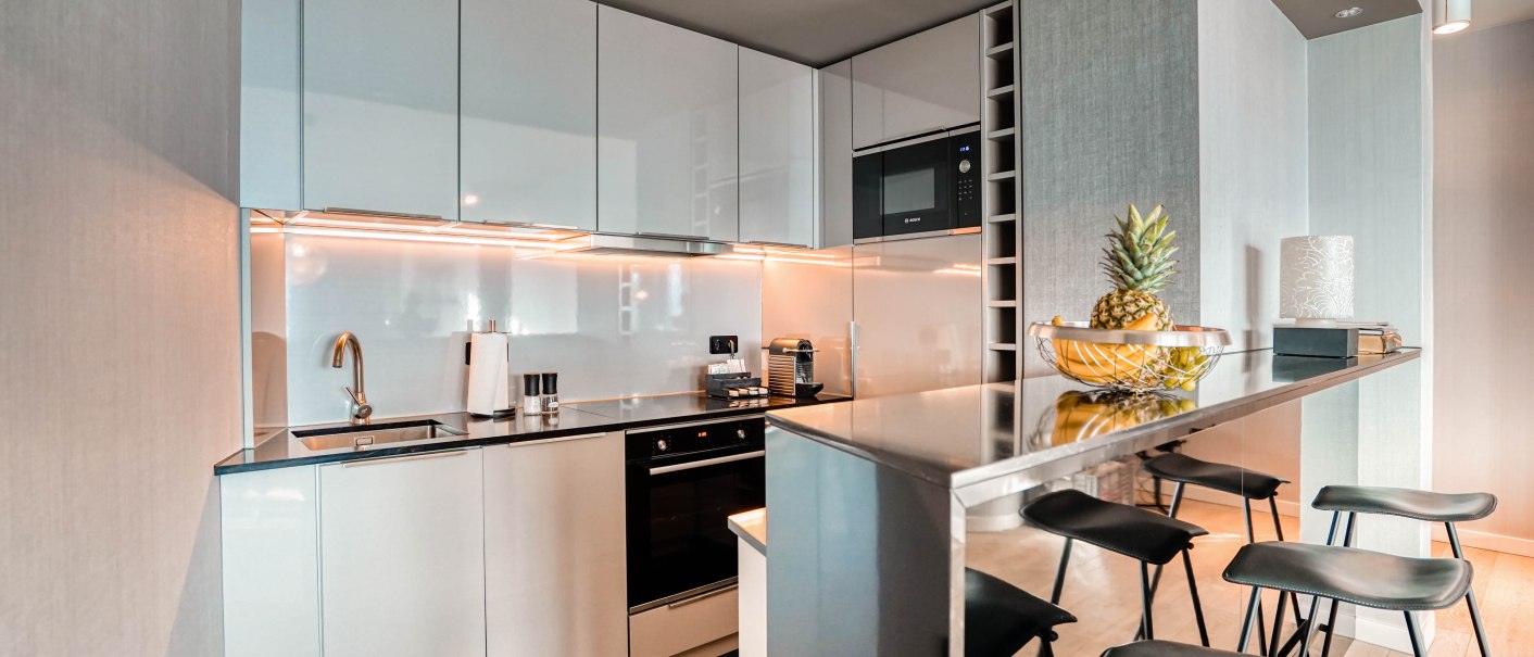 Moderne Küche mit weißen Hochglanzschränken, eingebautem Ofen und Mikrowelle. Eine Theke mit Barhockern und einer Obstschale mit Ananas., © CLOUD N°7 APARTMENTS GmbH Moderne Küche mit weißen Hochglanzschränken, eingebautem Ofen und Mikrowelle. Eine Theke mit Barhockern und einer Obstschale mit Ananas., © CLOUD N°7 APARTMENTS GmbH