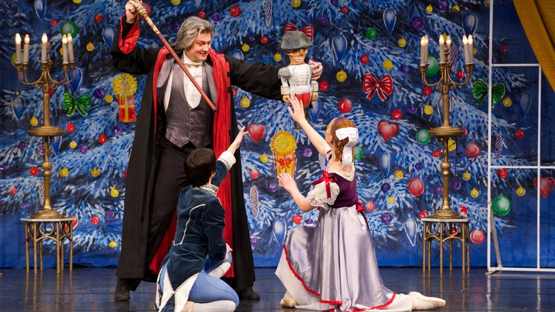 Ballettszene aus dem Nussknacker: Ein Mann in rotem Mantel hält eine Nussknackerfigur, während zwei Tänzer in historischen Kostümen knien. Hintergrund: Weihnachtsbaum., © Go 2 - Convent GmbH