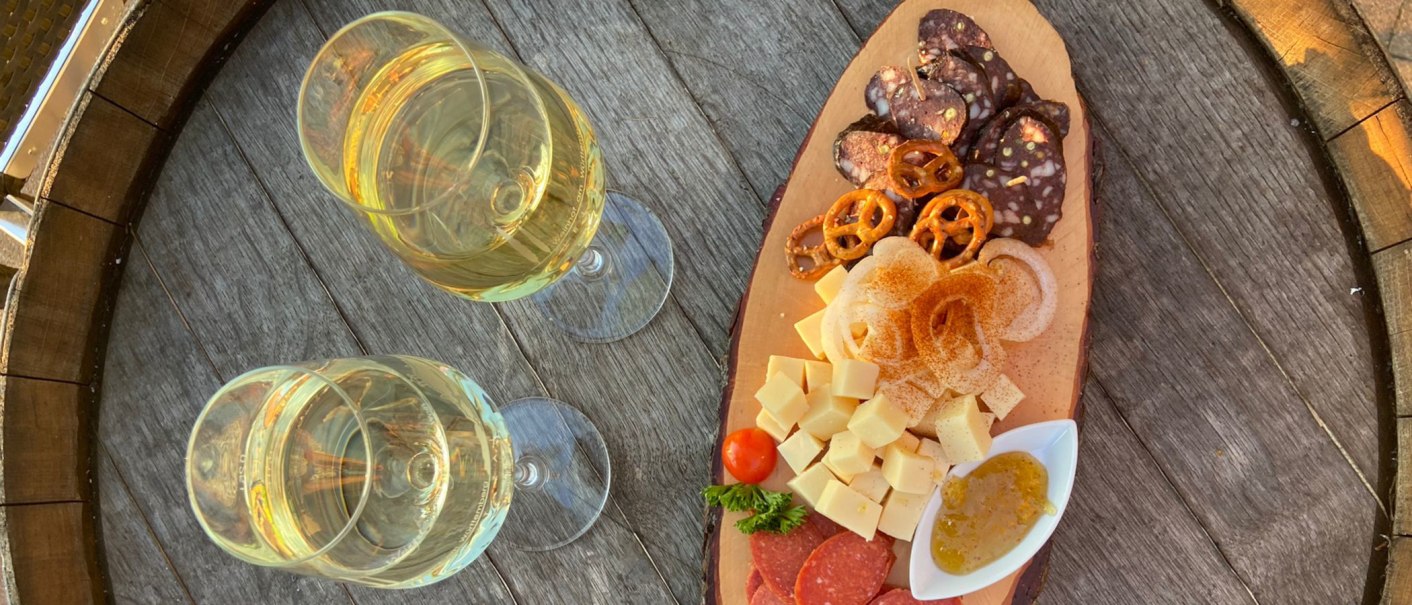 Zwei Gläser Weißwein neben einer Holzplatte mit Käsewürfeln, Wurst, Brezeln, Zwiebeln und Dip auf einem rustikalen Holztisch., © Weinhof Zaiß
