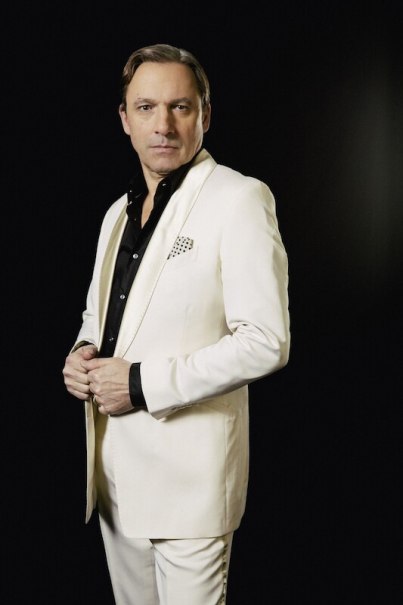 A man in an elegant white suit with a black shirt poses in front of a dark background., &copy; Theaterhaus Stuttgart e.V.