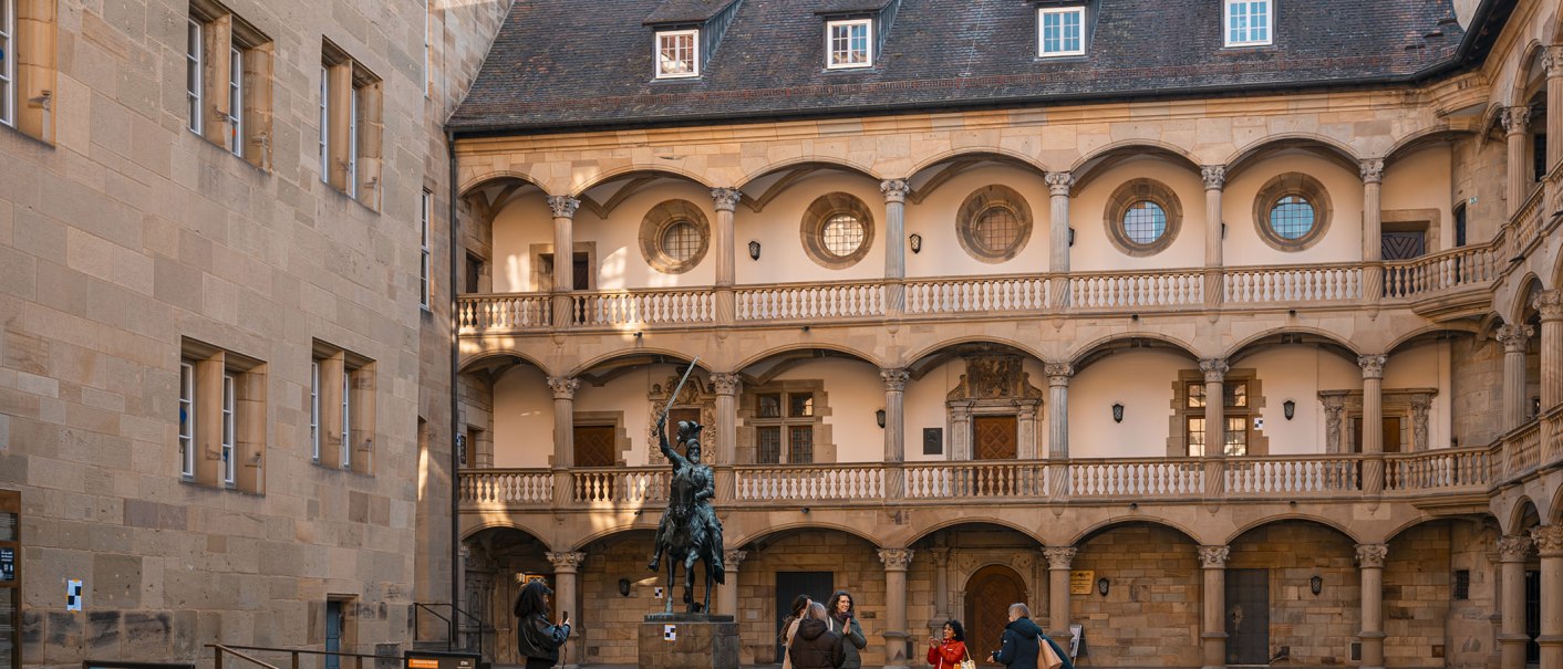Innenhof eines alten Schlosses mit Rundbögen, einer Reiterstatue und mehreren Personen, die Fotos machen. Historische Architektur und gepflasterter Boden., © Stuttgart Marketing GmbH - Sarah Schmid Innenhof eines alten Schlosses mit Rundbögen, einer Reiterstatue und mehreren Personen, die Fotos machen. Historische Architektur und gepflasterter Boden., © Stuttgart Marketing GmbH - Sarah Schmid