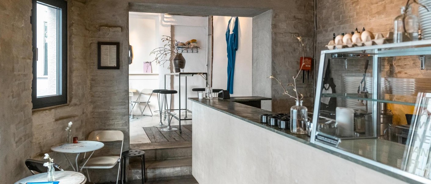 Modernes Café mit minimalistischer Einrichtung, weißen Wänden und Holzmöbeln. Ein blaues Kleidungsstück hängt an der Wand, Pflanzen auf Tischen., © SMG, Sarah Schmid