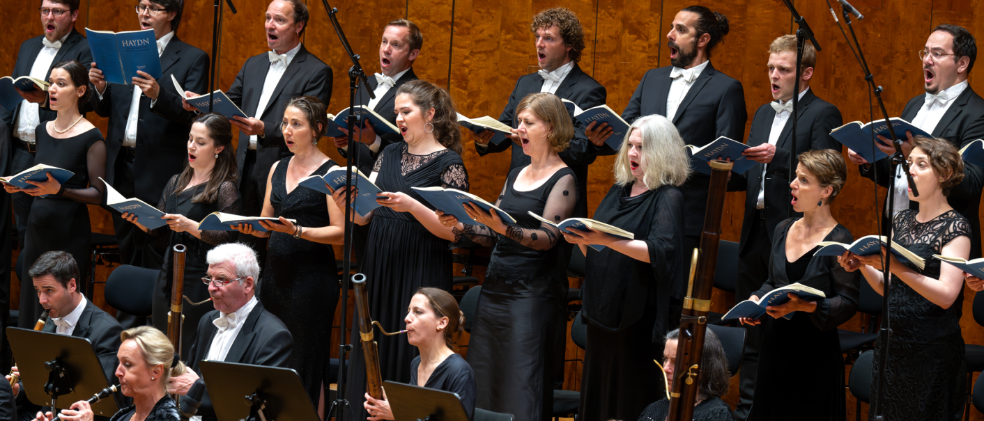 A choir and orchestra in elegant dress perform a work. The singers hold sheet music and sing with expression., &copy; Holger Schneider