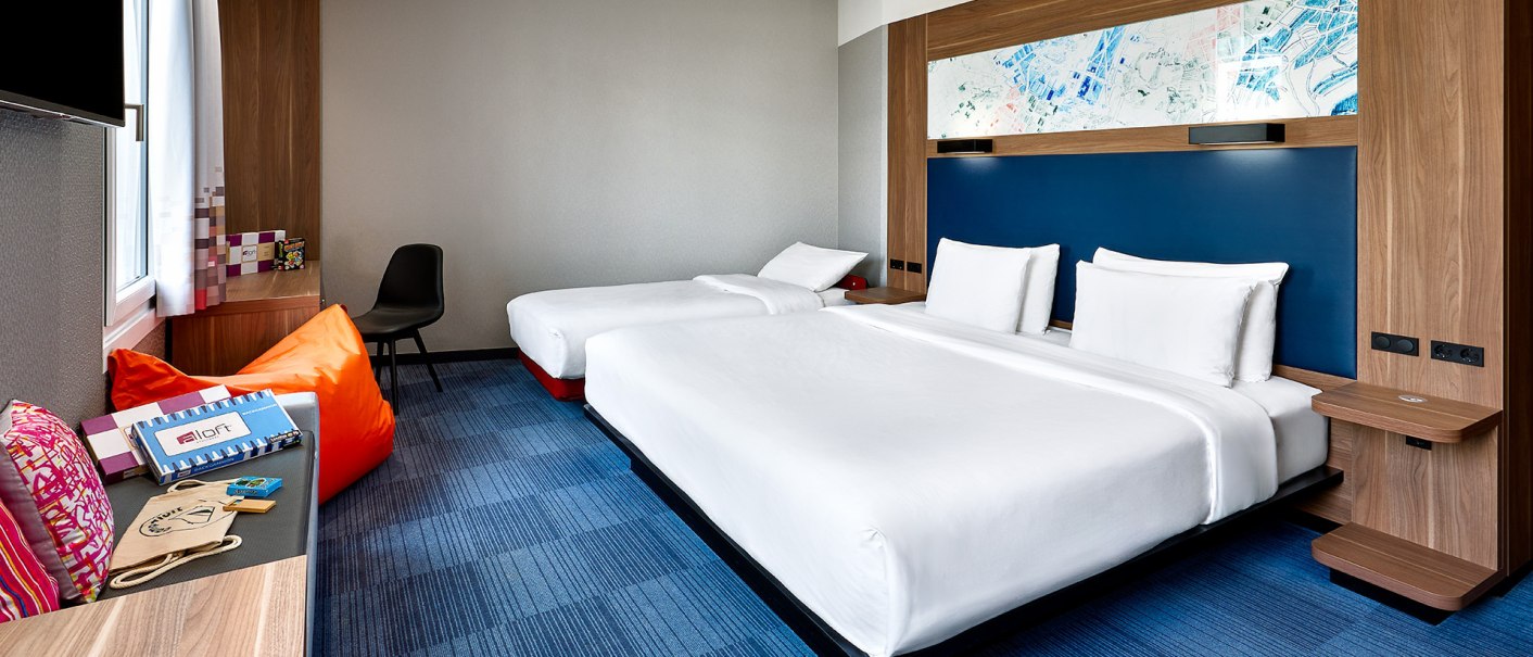 Modernes Hotelzimmer mit zwei Betten, einem Schreibtisch und einem Sitzbereich mit bunten Kissen. Helle, einladende Atmosphäre., © aloft Stuttgart Modernes Hotelzimmer mit zwei Betten, einem Schreibtisch und einem Sitzbereich mit bunten Kissen. Helle, einladende Atmosphäre., © aloft Stuttgart