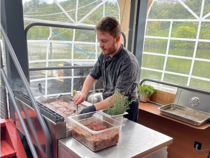 A cook grills meat on a boat. A river and green landscape can be seen in the background. Herbs and spices are ready., © Cool-Tours StattReisen A cook grills meat on a boat. A river and green landscape can be seen in the background. Herbs and spices are ready., © Cool-Tours StattReisen