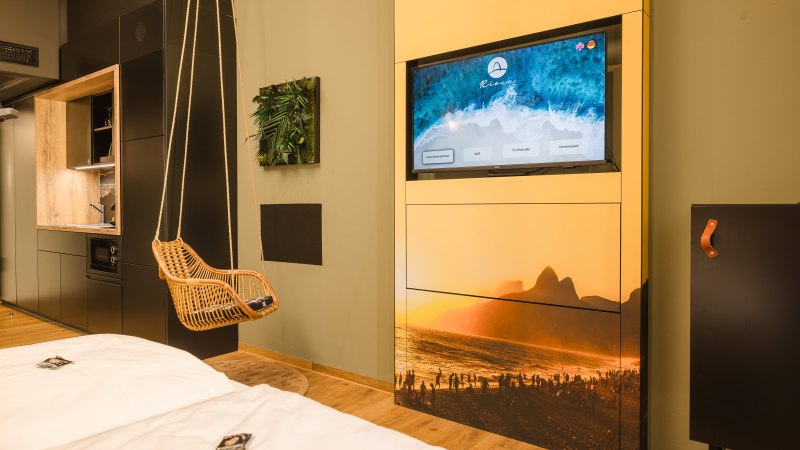 Modernes Hotelzimmer mit H&auml;ngesessel, Fernseher mit Strandbild, kleiner K&uuml;chenzeile und dekorativem Pflanzenbild an der Wand., &copy; TOMAS