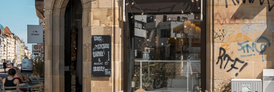 Café mit Außenbestuhlung, Graffiti an der Wand, Lichterketten über dem Eingang. Schild mit "Kaffee & Kuchen". Stadtstraße im Hintergrund., © SMG, Sarah Schmid Café mit Außenbestuhlung, Graffiti an der Wand, Lichterketten über dem Eingang. Schild mit "Kaffee & Kuchen". Stadtstraße im Hintergrund., © SMG, Sarah Schmid