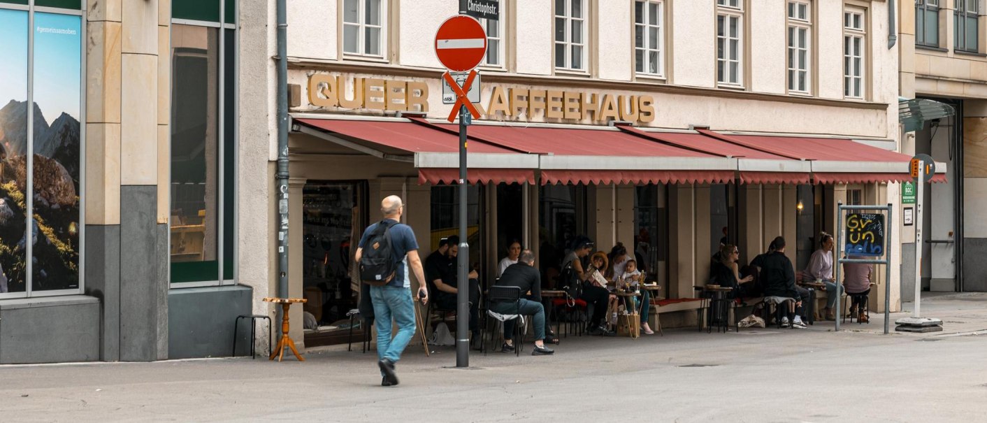 Ein Café mit rotem Vordach und dem Schriftzug 'Queer Kaffeehaus'. Menschen sitzen draußen an Tischen. Ein Mann geht vorbei., © SMG, Sarah Schmid Ein Café mit rotem Vordach und dem Schriftzug 'Queer Kaffeehaus'. Menschen sitzen draußen an Tischen. Ein Mann geht vorbei., © SMG, Sarah Schmid