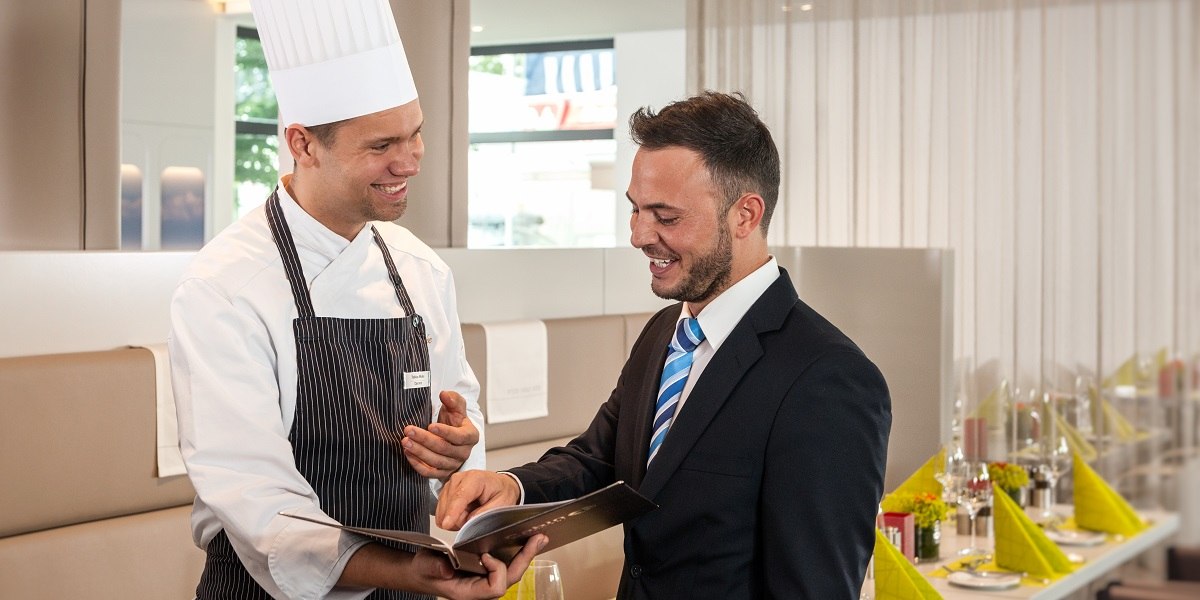Ein Koch in Uniform und ein Mann im Anzug l&auml;cheln und betrachten ein Buch in einem eleganten Restaurant., &copy; Essential by Dorint