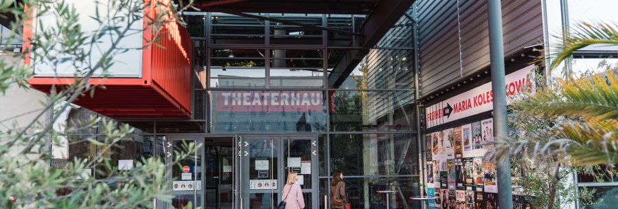 Entrance to the Theaterhaus Stuttgart with red floor, glass facade and posters. Two people enter the building. Plants and modern architecture are visible., © Stuttgart-Marketing GmbH, wpsteinheisser