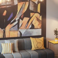 A modern lobby with a gray sofa, colorful pillows, an abstract wall hanging, a yellow side table, a plant, and a floor lamp., © the niu Form - Novum Hospitality A modern lobby with a gray sofa, colorful pillows, an abstract wall hanging, a yellow side table, a plant, and a floor lamp., © the niu Form - Novum Hospitality