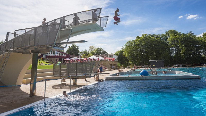 Ein Freibad mit Sprungturm, von dem ein Mensch ins Wasser springt. Viele Menschen schwimmen im gro&szlig;en Becken. Im Hintergrund sind B&auml;ume und Sonnenschirme., &copy; Stuttgarter B&auml;der