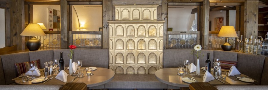 Cozy restaurant corner with wooden tables, covered seating and a decorative tiled stove in the middle., © Hotel Gloria GmbH