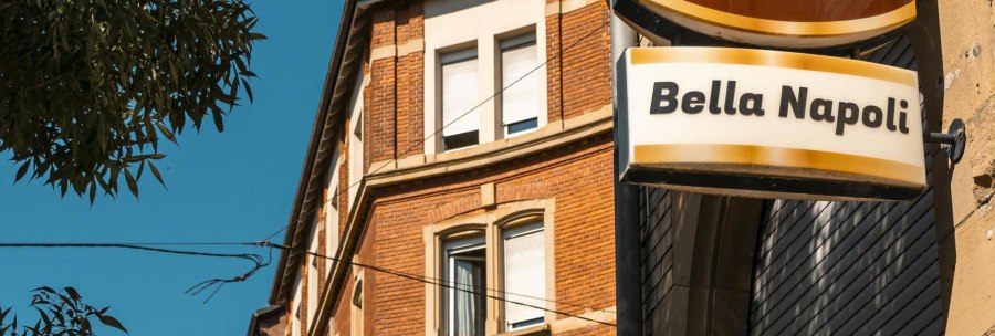 Ein Schild mit der Aufschrift 'Bella Napoli' h&auml;ngt an einem Backsteingeb&auml;ude. Der Himmel ist klar und blau, ein Baum ist im Vordergrund sichtbar., &copy; SMG, Sarah Schmid