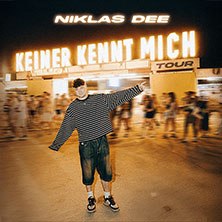 Eine Person posiert vor einem beleuchteten Schild mit der Aufschrift 'Keiner kennt mich Tour'. Die Umgebung ist belebt und unscharf., &copy; links im Bild