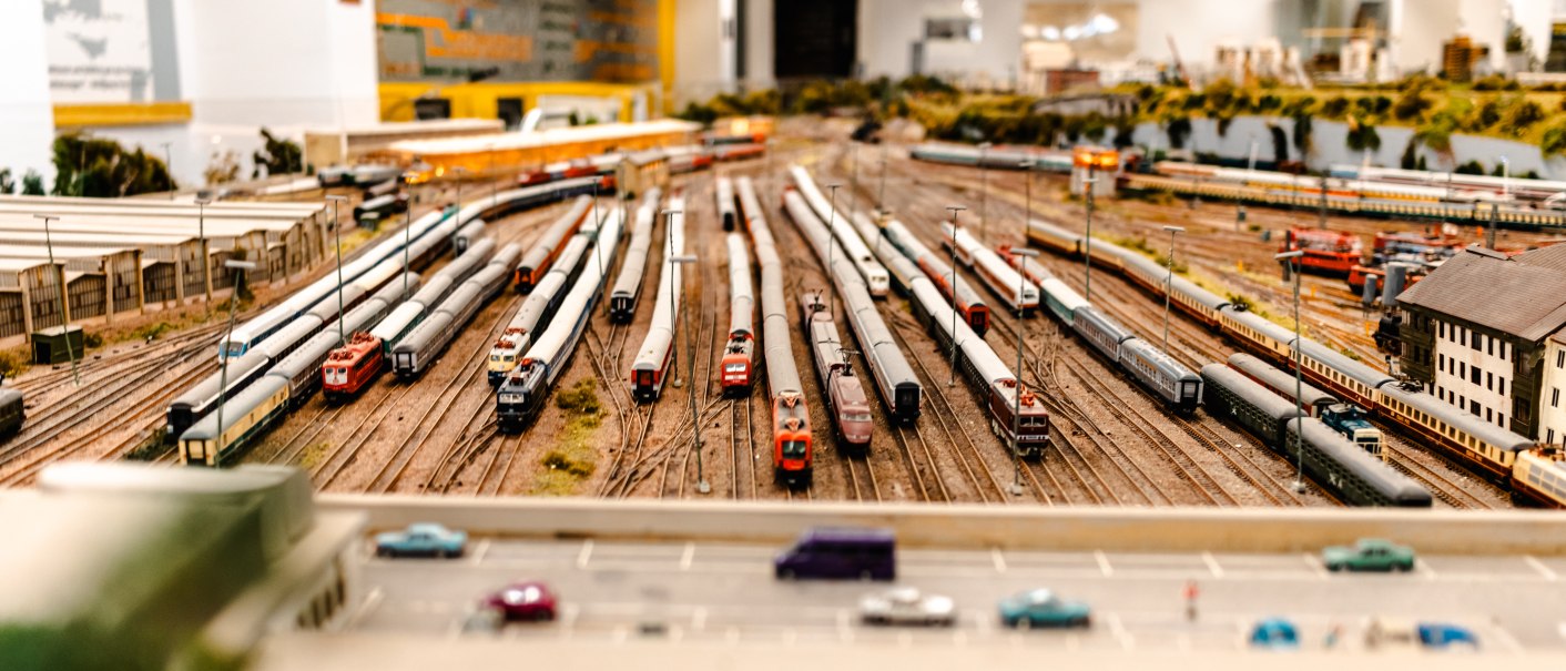 Miniatur-Eisenbahnanlage mit zahlreichen Zügen auf Gleisen in einer Ausstellung. Im Vordergrund sind Autos auf einer Straße zu sehen., © SMG_Sarah Schmid