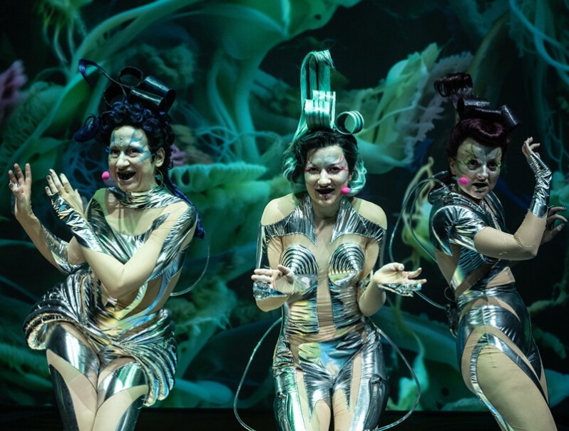 Three performers in silver, futuristic costumes perform on a stage in front of a green, abstract background., © Württembergische Staatstheater Stuttgart Three performers in silver, futuristic costumes perform on a stage in front of a green, abstract background., © Württembergische Staatstheater Stuttgart